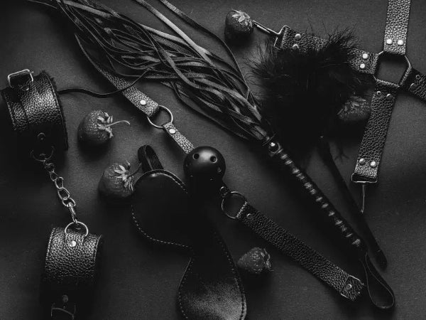 LaceBound black leather BDSM kit flatlay featuring blindfold, wrist cuffs, and whip arranged on neutral background for sensual accessory display